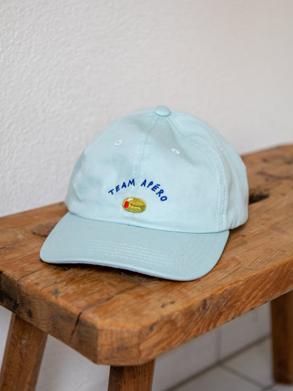 Mint farbenes Dad Cap mit der Stickerei Team Apéro und einer gestickten Olive auf einer urchigen Bank mit weissem Hintergrund.