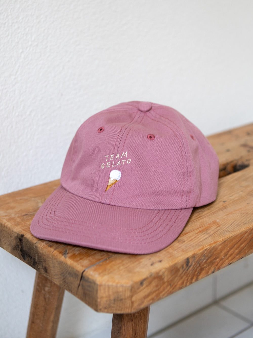 Rosa farbenes Dad Cap mit der Stickerei Team Gelato und einem Glacé in der Waffel auf einer urchigen Bank mit weissem Hintergrund.