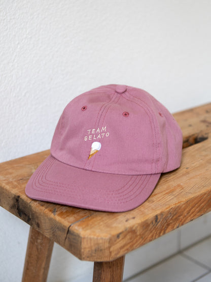 Rosa farbenes Dad Cap mit der Stickerei Team Gelato und einem Glacé in der Waffel auf einer urchigen Bank mit weissem Hintergrund.