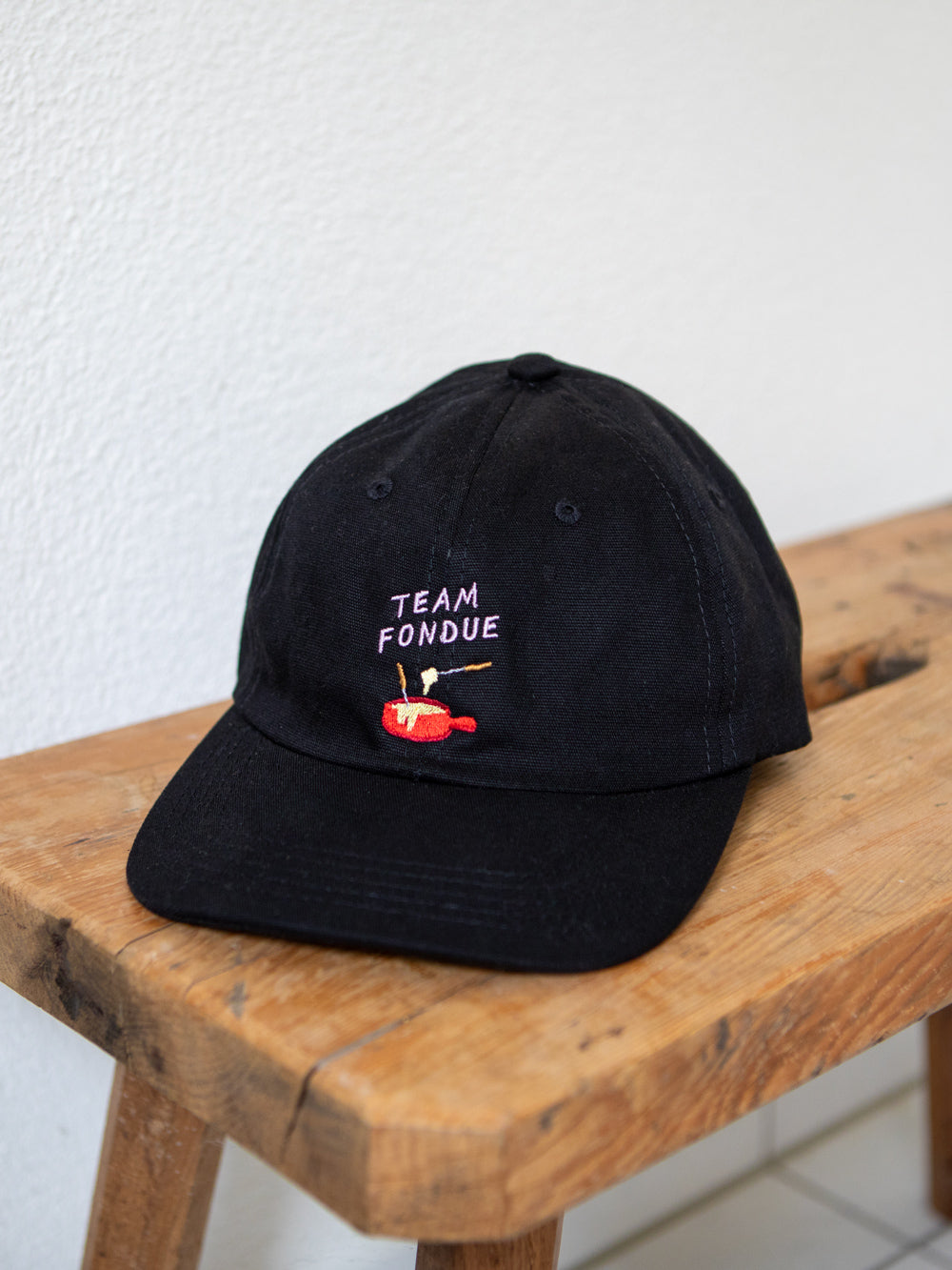 Schwarzes Dad Cap liegend auf brauner rustikaler Bank mit blauem gesticktem Schriftzug "Team Fondue" sowie rotem gesticktem Fondue Caquelon.