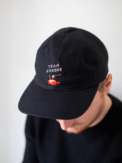 Oberkörper eines Mannes, der ein schwarzes Cap trägt. Darauf ist eine Stickerei mit dem Schriftzug Team Fondue und der Abbildung eines roten Fondue Caquelons.