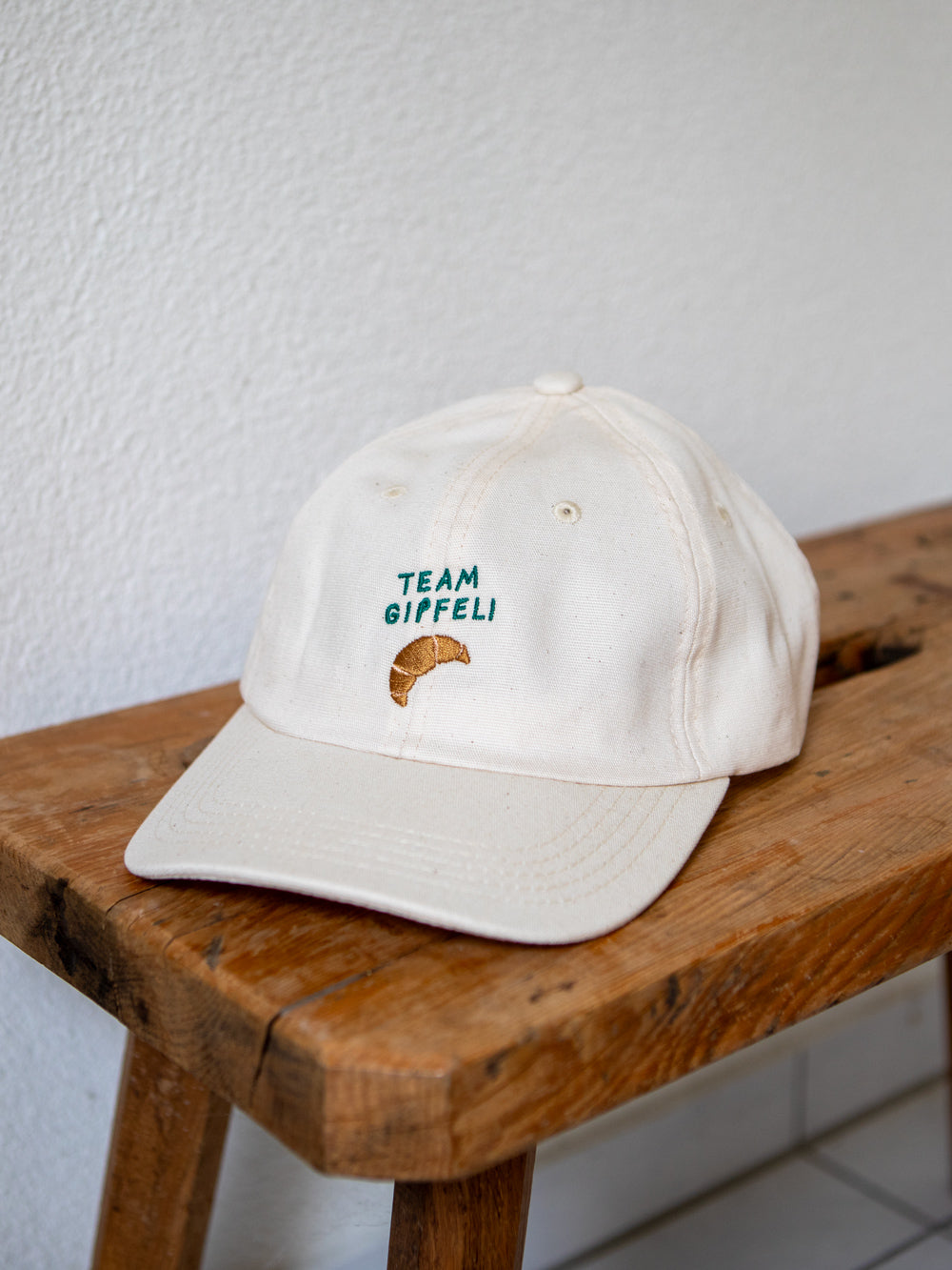 Ecru farbenes Cap mit grünem gesticktem Schriftzug Team Gipfeli und einem gestickten Croissant liegt auf einem rustikalen Holzbank.