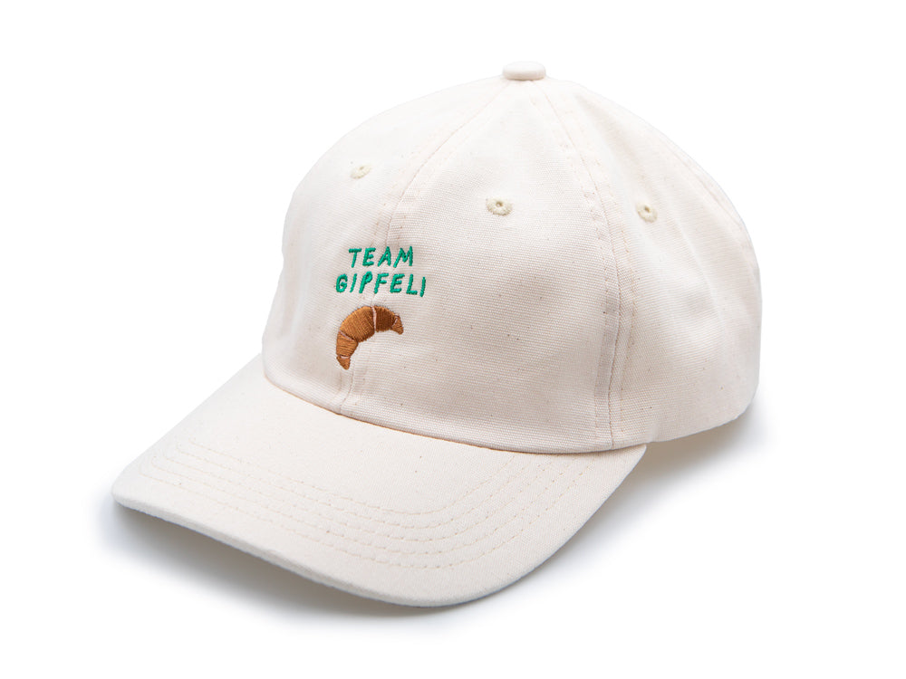 Ecru farbenes Cap mit grünem gesticktem Schriftzug Team Gipfeli und einem gestickten Croissant auf weissem Hintergrund.