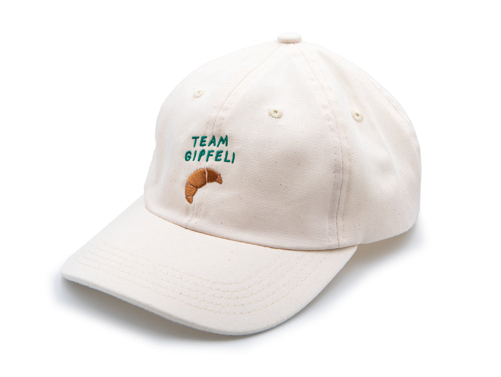 Ecru farbenes Cap mit grünem gesticktem Schriftzug Team Gipfeli und einem gestickten Croissant auf weissem Hintergrund.
