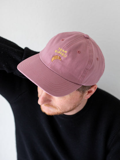 Oberkörper eines Mannes, der ein rosa farbenes Cap trägt. Darauf ist eine Stickerei mit dem gelben Schriftzug Team Gipfeli und der Croissants.