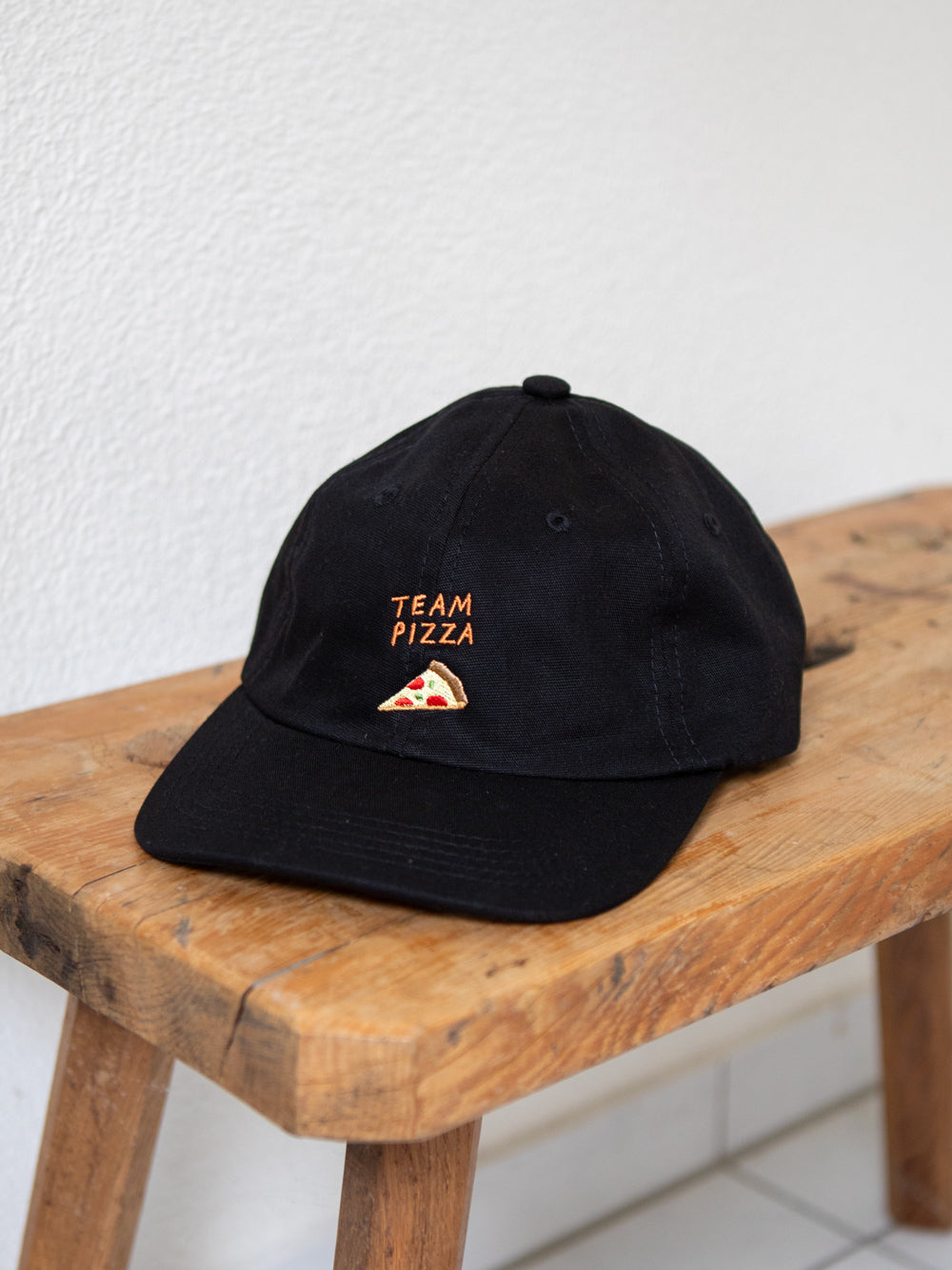 Schwarzes Dad Cap mit der Stickerei Team Pizza und einem gestickten Stück Pizza auf einer urchigen Bank mit weissem Hintergrund.