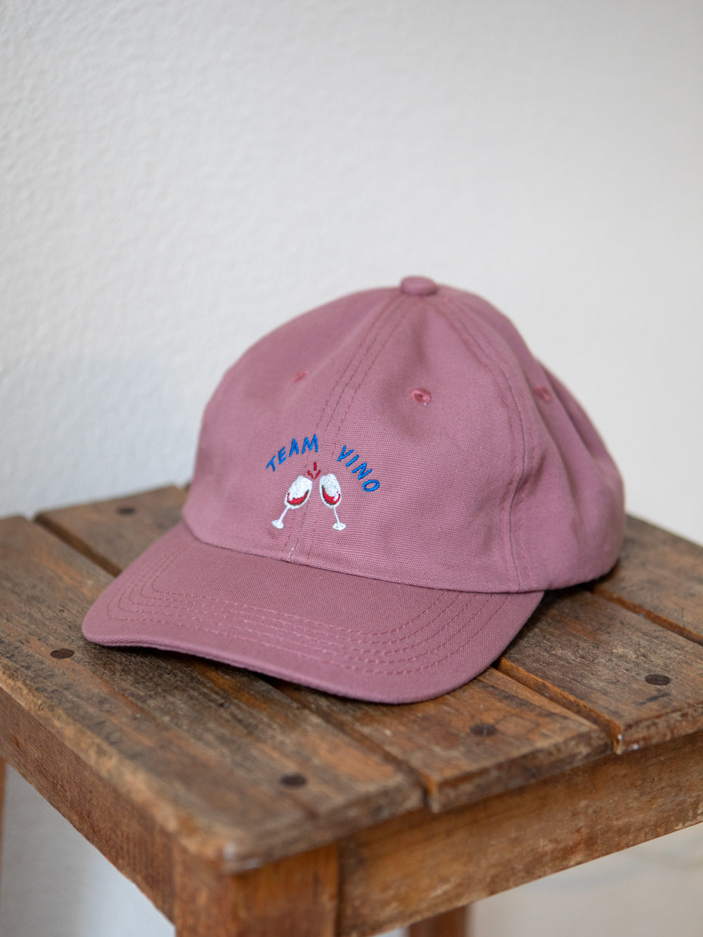 Rosa farbenes Cap mit gesticktem Schriftzug Team Vino und zwei gestickten Weingläsern auf rustikalem Hocker.