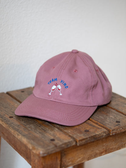 Rosa farbenes Cap mit gesticktem Schriftzug Team Vino und zwei gestickten Weingläsern auf rustikalem Hocker.