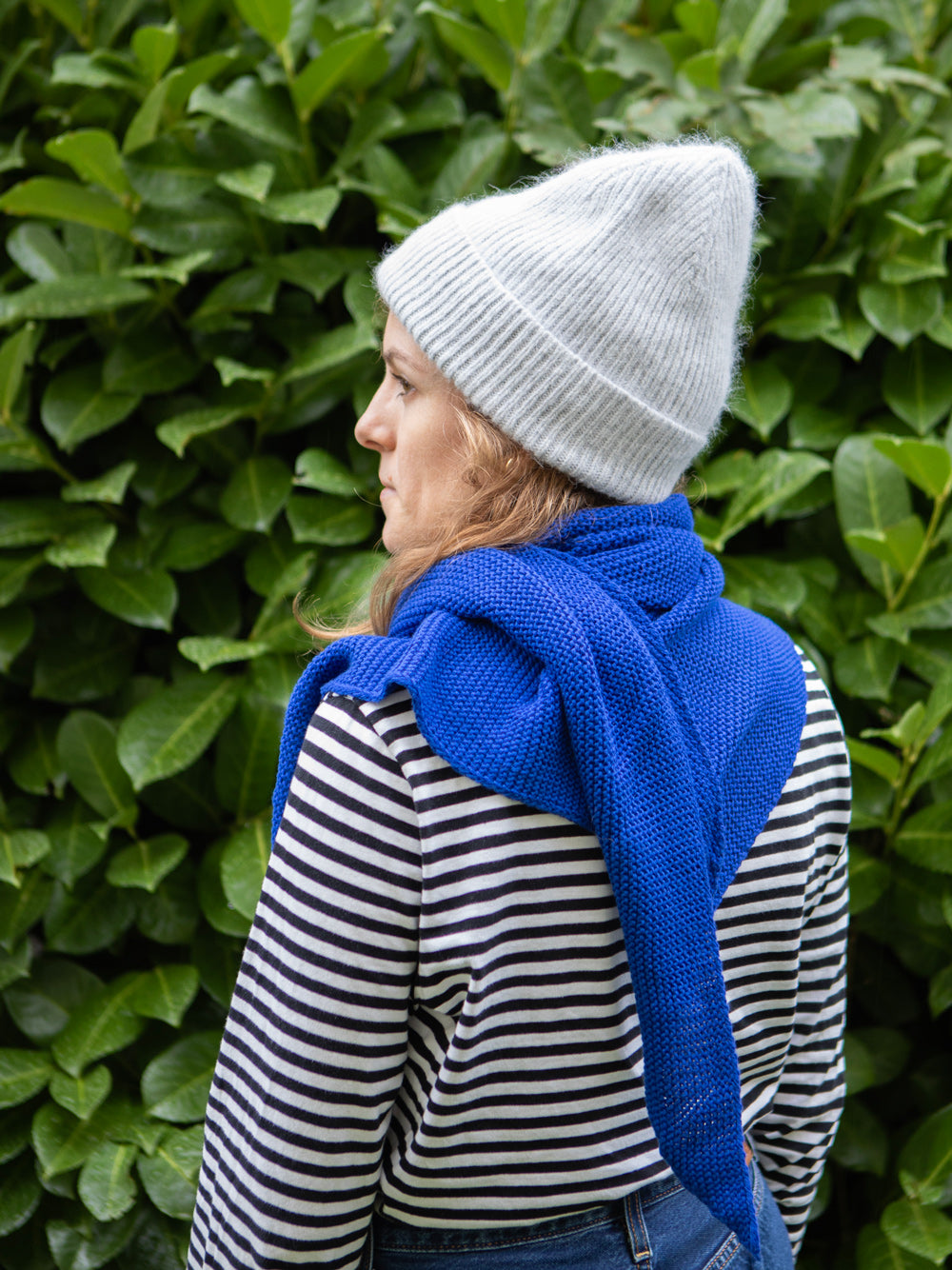 Bild einer Frau von hinten mit grauer Wollmütze und blauem gestricktem Schal über einem gestreiften Shirt. Im Hintergrund sieht man unscharfe Blätter.