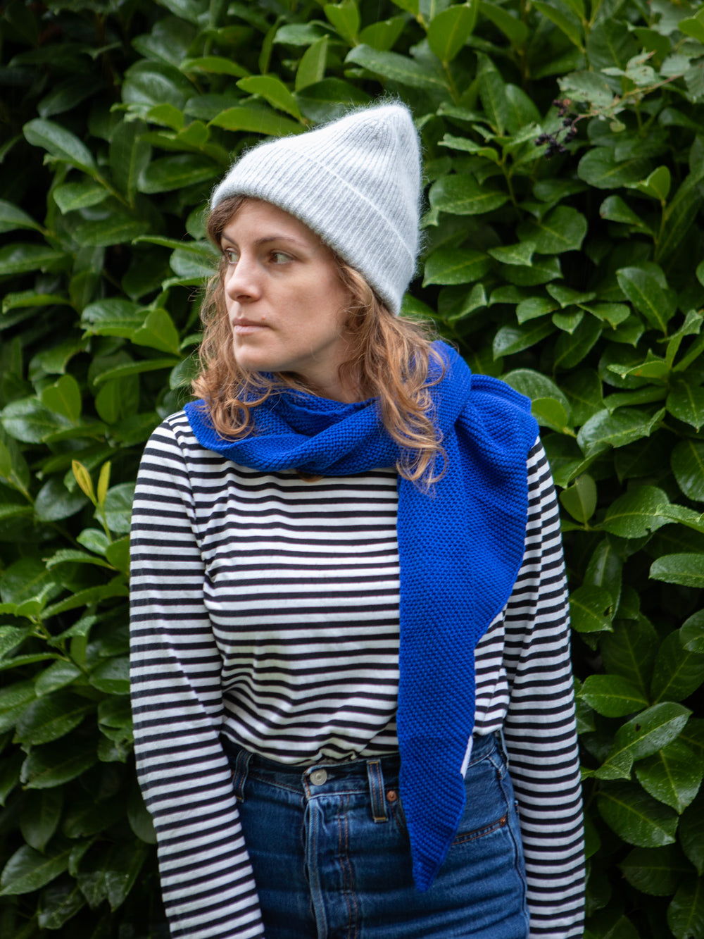 Frau mit langen braunen Haaren trägt eine flauschige hellgraue Wollmütze, ein gestreiftes Shirt und einen blauen gestrickten Schal. Im Hintergrund sieht man unscharfe Blätter.