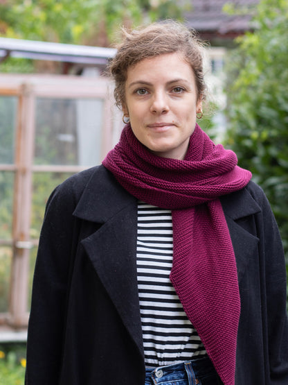 Frau mit braunen Haaren trägt einen gestrickten bordeaux farbenen Dreiecksschal über einem gestreiften Shirt mit schwarzer Jacke. Unscharfer Hintergrund.