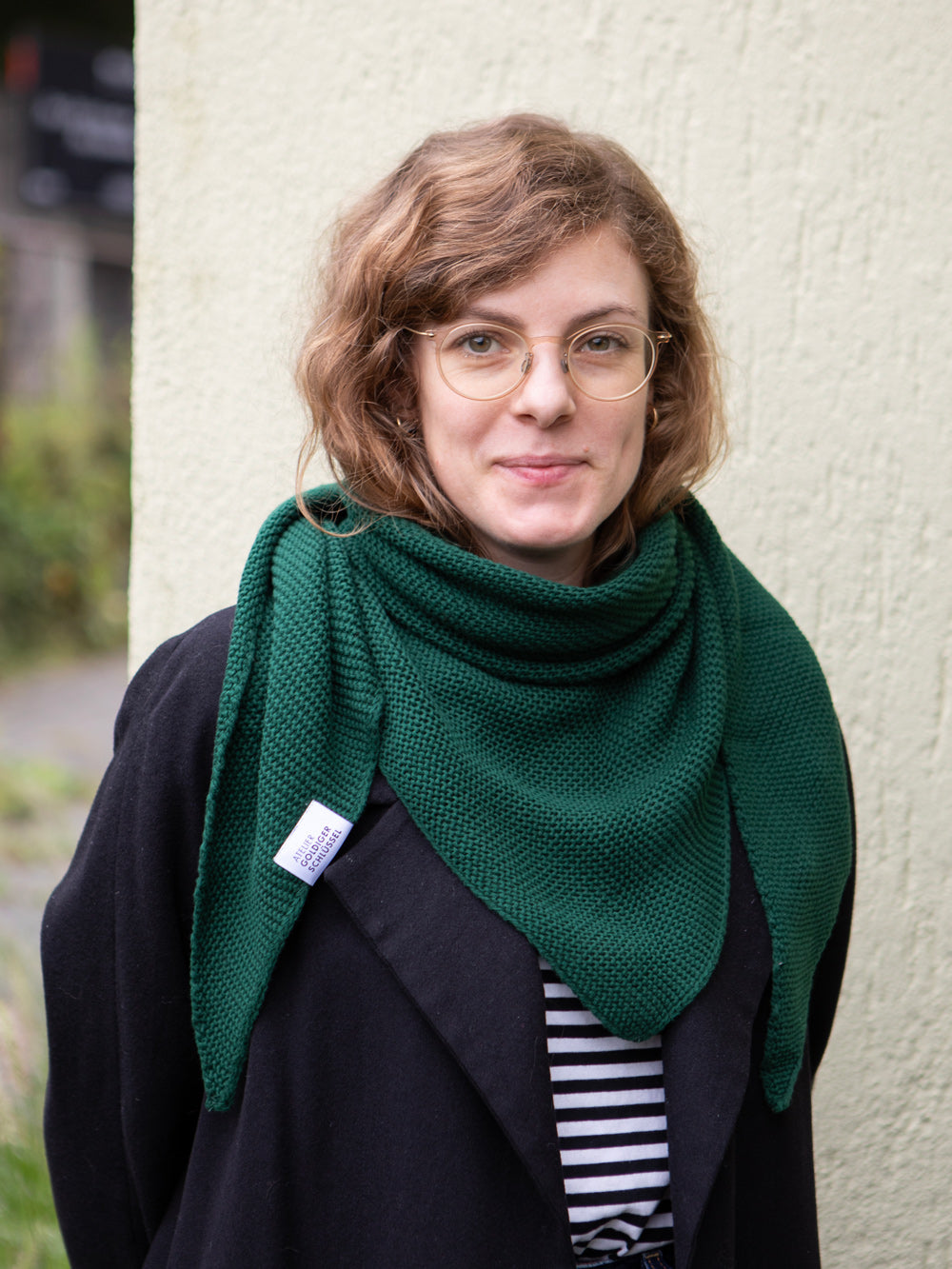 Frau mit blondbraun gelockten Haaren und Brille trägt einen gestrickten dunkelgrünen Dreiecksschal über einer schwarzen Wolljacke. Im Hintergrund eine unscharfe Fassade.
