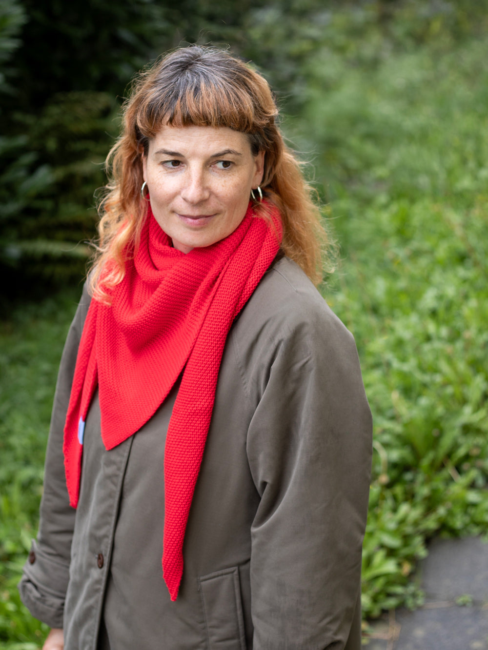 Frau mit orange-braunen gelockten Haaren trägt einen gestrickten roten Dreiecksschal und eine olivgrüne Jacke. Unscharfe Wiese im Hintergrund.