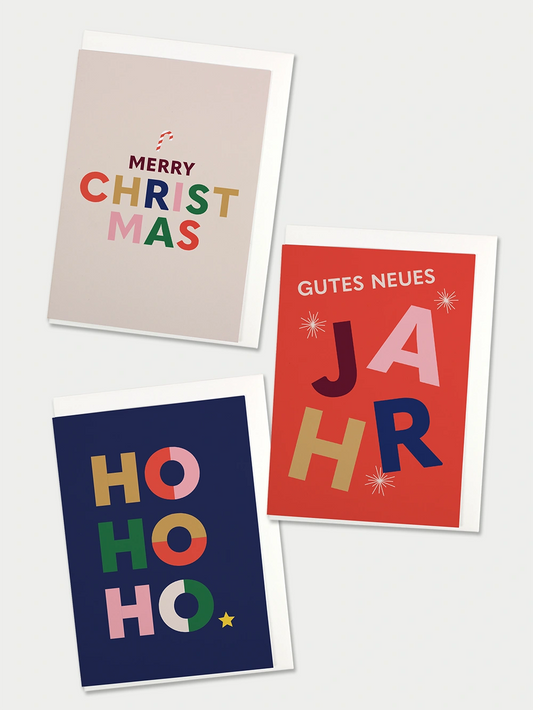 Drei Faltkarten mit Couvert, eine beige mit farbigem Schriftzug Merry Christmas, eine dunkelblau mit Schriftzug HOHOHO, eine rot mit dem Schriftzug Gutes Neues Jahr