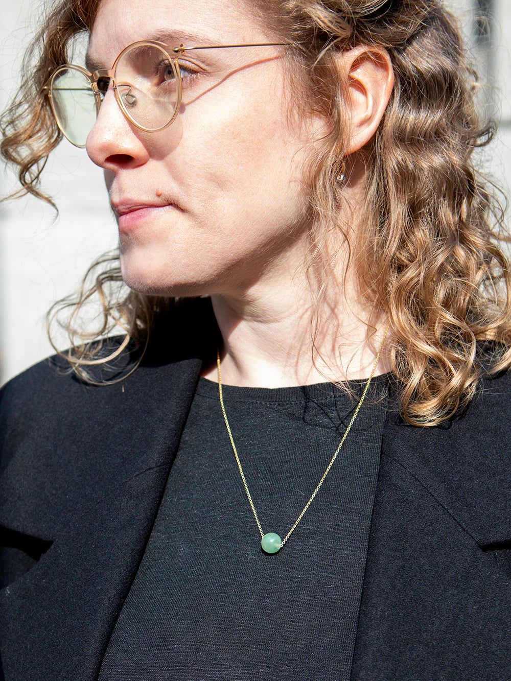 Oberkörper eine Frau mit lockigen Haaren und einer transparenten Brille welche eine goldene Kette mit einer Perle aus türkisfarbenem Aventurin trägt.