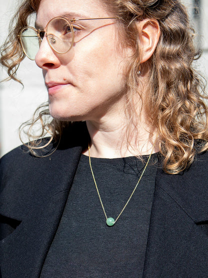 Oberkörper eine Frau mit lockigen Haaren und einer transparenten Brille welche eine goldene Kette mit einer Perle aus türkisfarbenem Aventurin trägt.