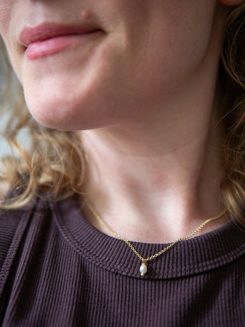 Detailaufnahme einer Person die eine goldene Halskette mit einer weissen Süsswasserperle als Anhänger über einem braun gerippten Shirt trägt.
