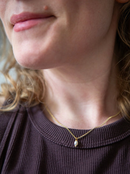 Detailaufnahme einer Person die eine goldene Halskette mit einer weissen Süsswasserperle als Anhänger über einem braun gerippten Shirt trägt.