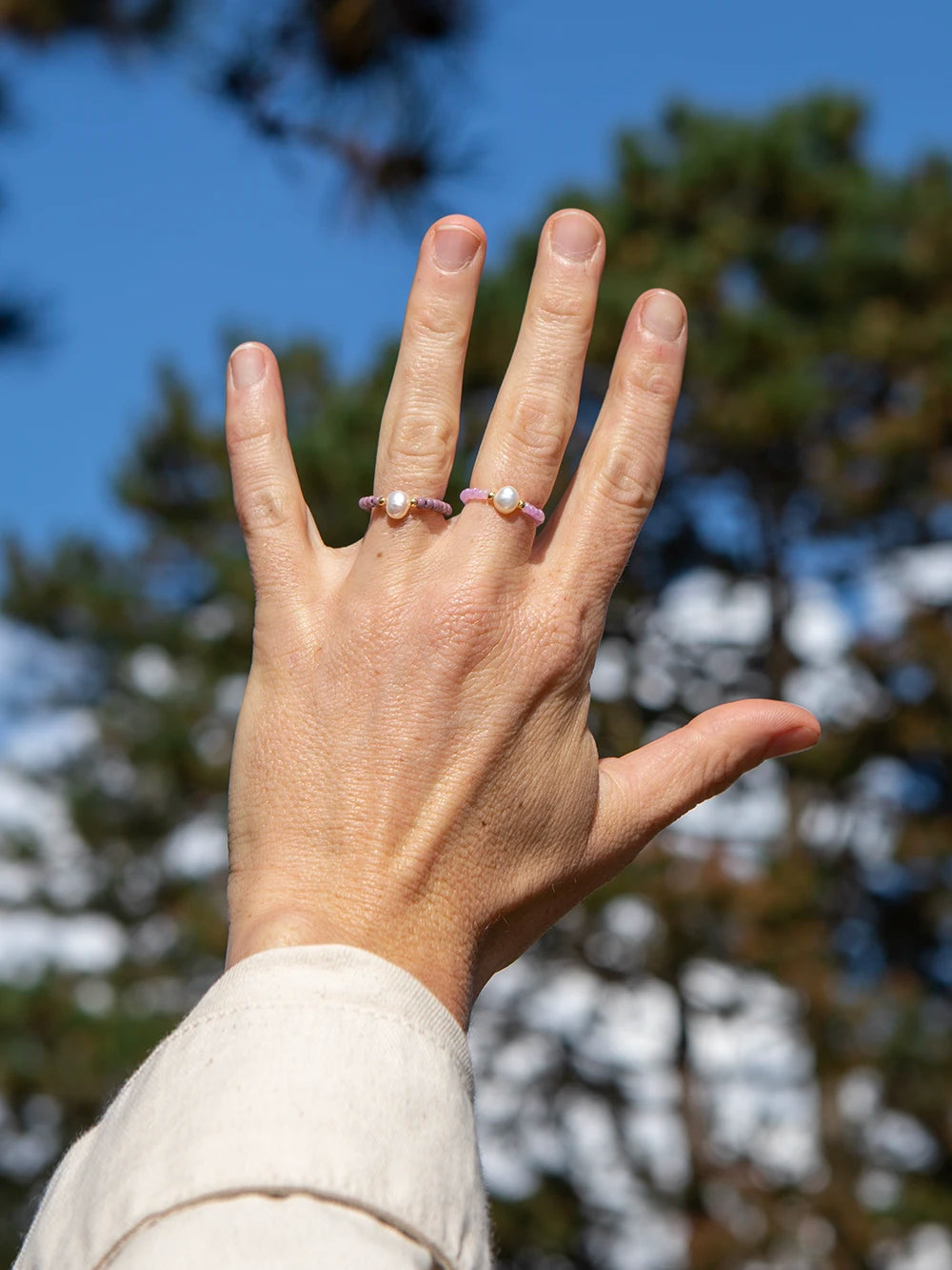 Detailansicht einer Hand, welche zwei rosa und lila farbene Ringe mit je einer Süsswasserperle trägt. Im Hintergrund sieht man unscharf einen Himmel und Bäume.