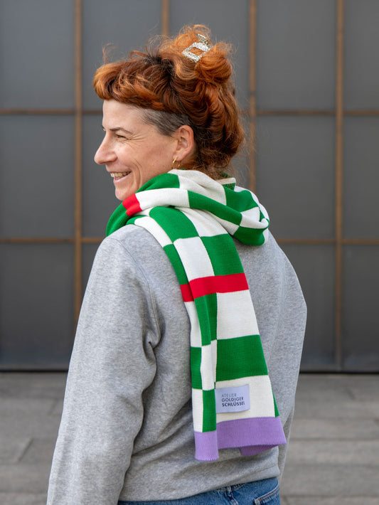 Frau mit orangen Haaren trägt grün-weiss karierten Schal über grauem Pullover. Blick nach links aus dem Bild.