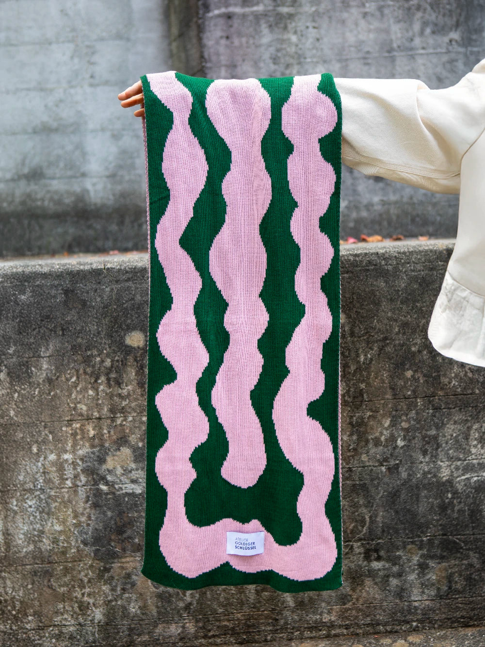 Ein Arm hält von rechts einen grün rosa gemusterten Strickschal ins Bild.