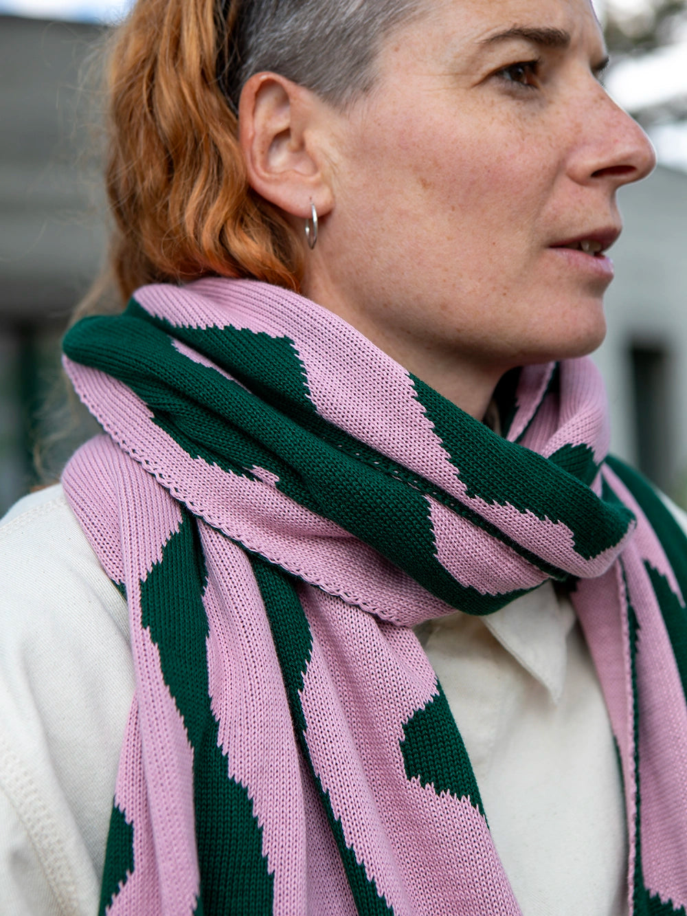 Nahaufnahme einer Frau mit orangen Haaren die einen grün-rosa gemusterten Strickschal und eine Jacke in offwhite trägt.