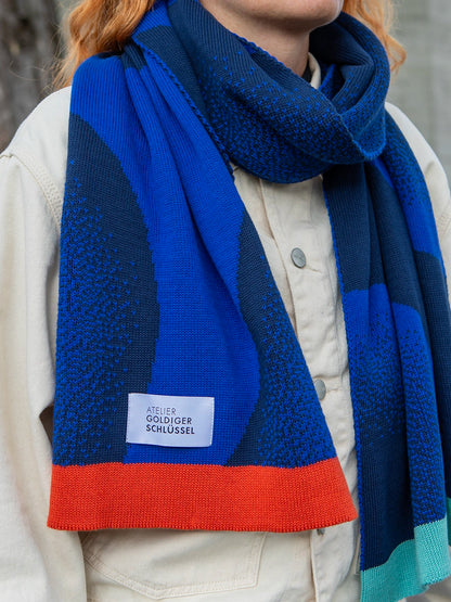 Detailaufnahme einer Person die einen gestrickten Schal in zwei unterschiedlichen Blautönten und mit bunten Abschlüssel trägt.