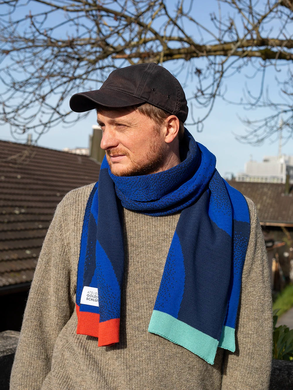 Mann mit schwarzem Cap und beigen Wollpullover trägt einen blau gemusterten Schal von Atelier Goldiger Schlüssel mit bunten Enden in mint und rostorange.