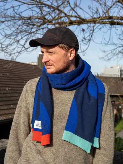 Mann mit schwarzem Cap und beigen Wollpullover trägt einen blau gemusterten Schal von Atelier Goldiger Schlüssel mit bunten Enden in mint und rostorange.