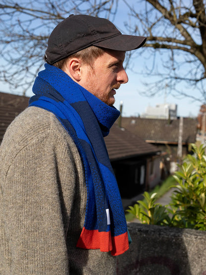Mann mit schwarzem Cap und beigen Wollpullover trägt einen blau gemusterten Schal von Atelier Goldiger Schlüssel mit bunten Enden in mint und rostorange.