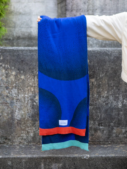 Arm hält von rechts einen gestrickten Schal in zwei unterschiedlichen Blautönen und mit bunten Abschlüssen ins Bild.