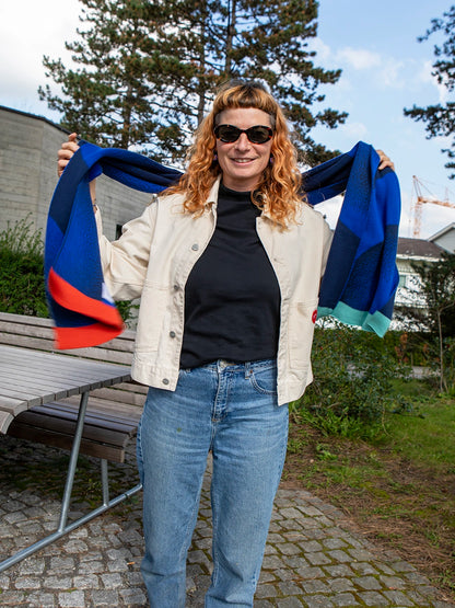 Stehende Frau mit orangen Haaren und Sonnenbrille trägt einen Schal in zwei unterschiedlichen Blautönen und mit bunten Abschlüssen.