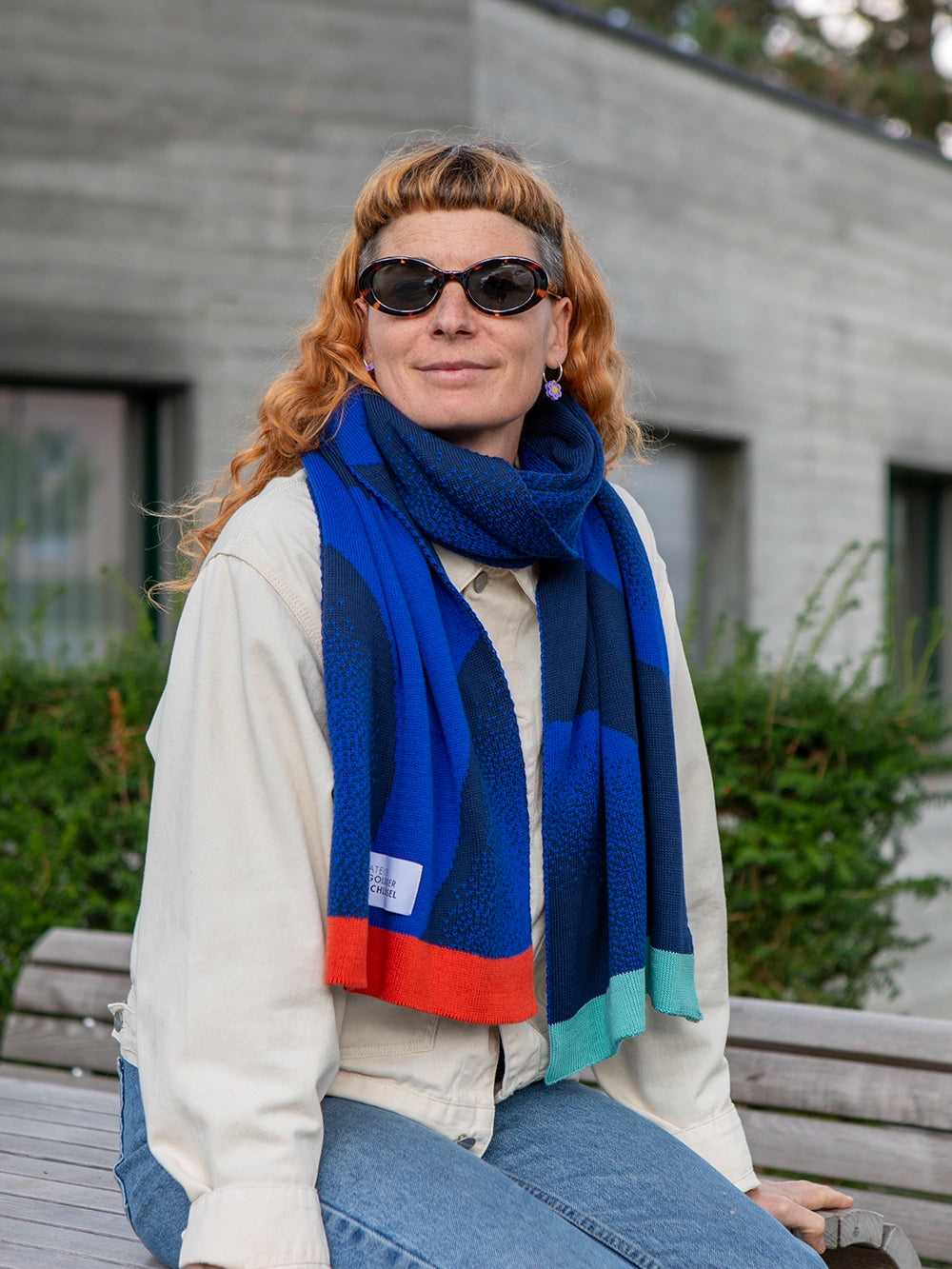 Sitzende Frau mit orangen Haaren und Sonnenbrille trägt einen Schal in zwei unterschiedlichen Blautönen und mit bunten Abschlüssen.