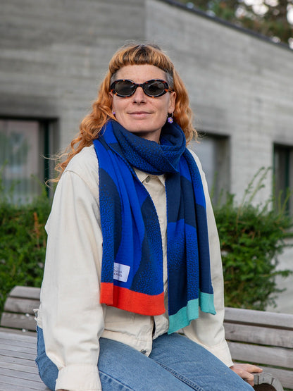 Sitzende Frau mit orangen Haaren und Sonnenbrille trägt einen Schal in zwei unterschiedlichen Blautönen und mit bunten Abschlüssen.