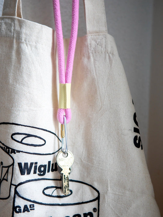 Detailaufnahme von einem rosa Schlüsselband aus Baumwollkordel mit silbernem Karabiner. Im Hintergrund ist eine Tote Bag angeschnitten.