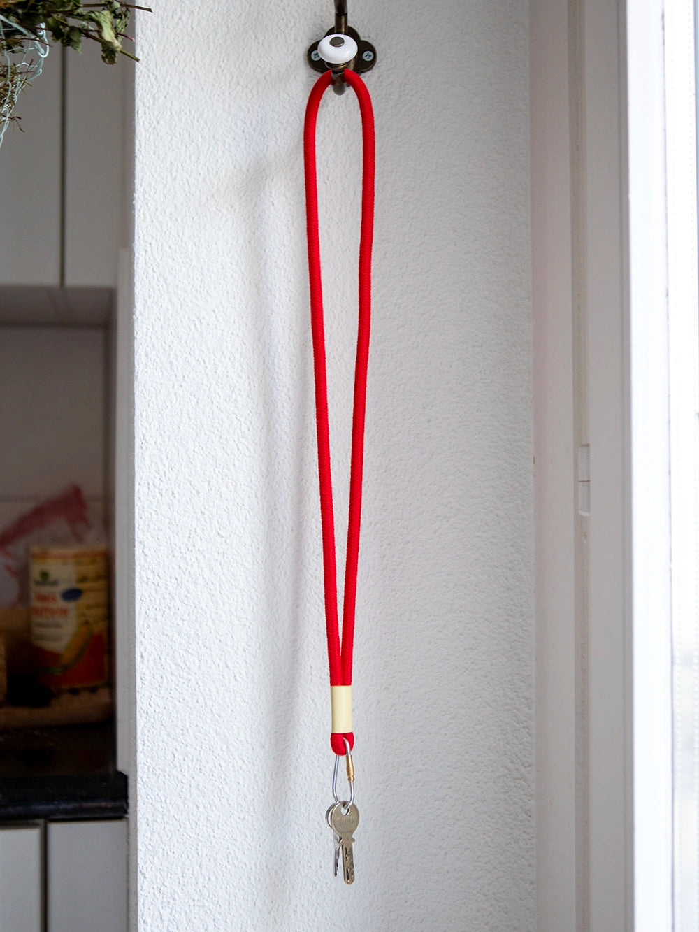 Rotes Schlüsselband aus Baumwollkordel mit silbernem Karabiner hänge an Haken vor weisser Wand.