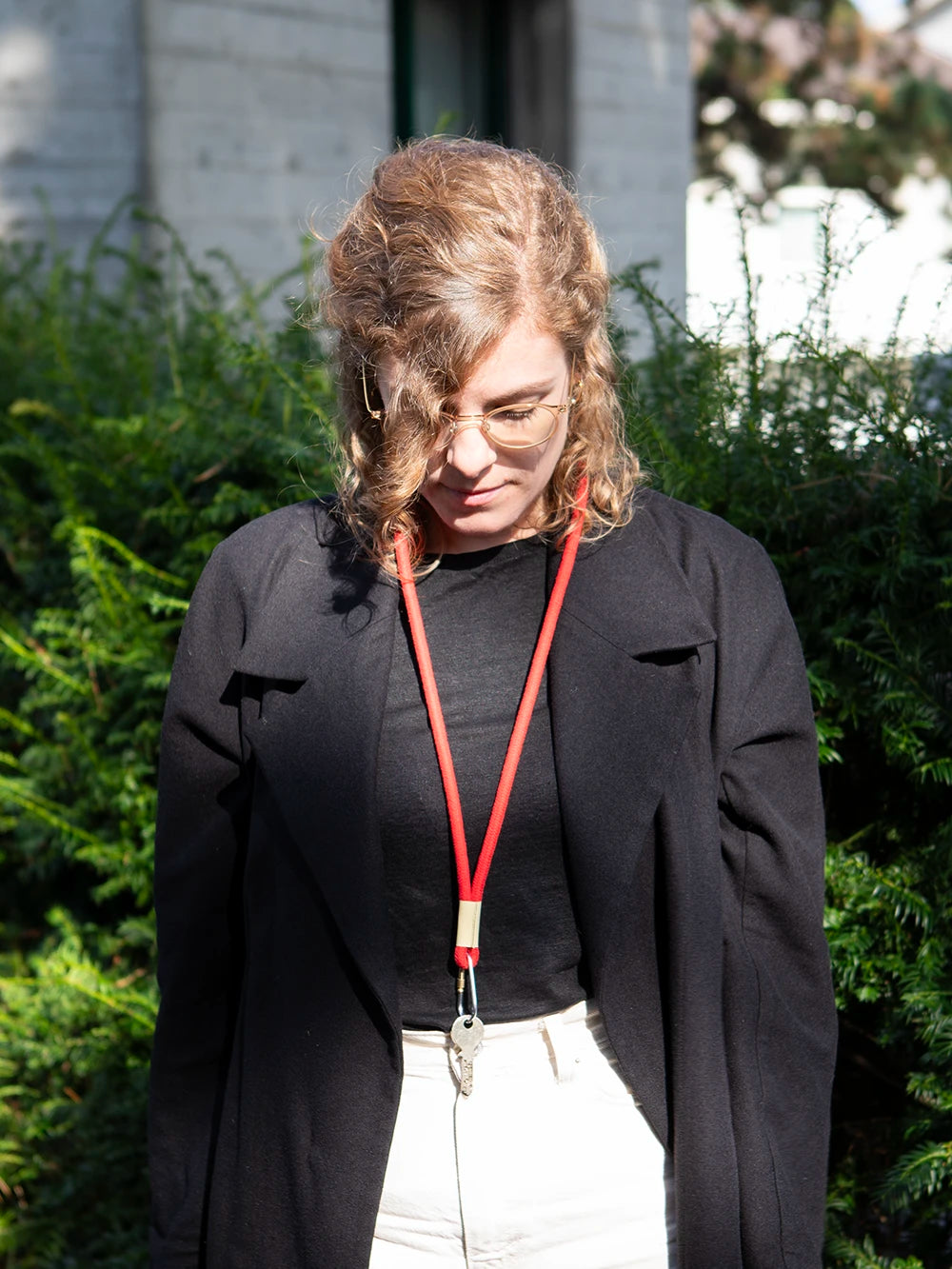 Oberkörper einer Frau mit blonden lockigen Haaren die ein schwarzes Oberteil und ein rotes Schlüsselband um den Hals trägt. Im Hintergrund sieht man unscharf grüne Büsche und ein Gebäude.