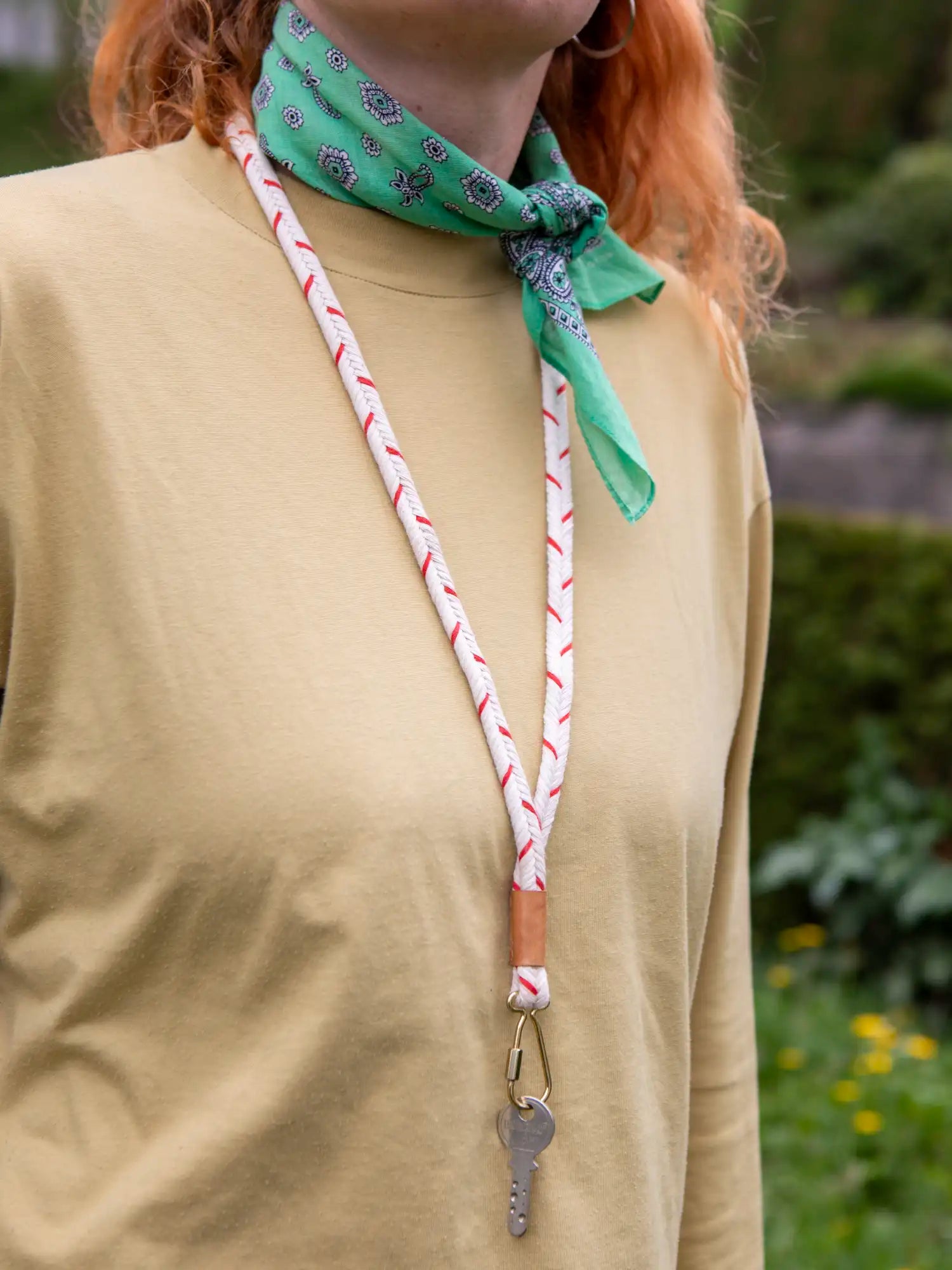 Oberkörper einer Frau mit orangen Haaren mit einem gelblichen Shirt und einem grünen Halstuch. Um den Hals trägt die Person ein rot weiss gemustertes und geflochtenes Schlüsselband mit einem Schlüssel an goldenem Karabiner.