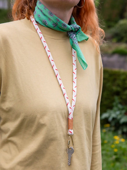 Oberkörper einer Frau mit orangen Haaren mit einem gelblichen Shirt und einem grünen Halstuch. Um den Hals trägt die Person ein rot weiss gemustertes und geflochtenes Schlüsselband mit einem Schlüssel an goldenem Karabiner.