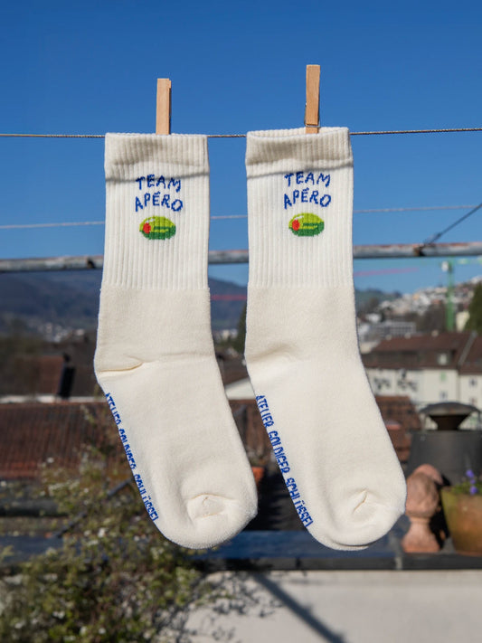 Zwei Sportsocken in ecru hängen an einer Wäscheleine, im Hintergrund unscharfe Häuser und Hügel. Im Bündchen wurde der blaue Schriftzug Team Apéro sowie eine grüne Olive eingestrickt.
