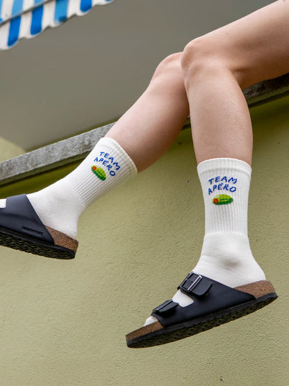 Beine über Brüstung die schwarze Birkenstock Arizona Sandalen sowie ecru farbene Team Apéro Sportsocken tragen. Am oberen Bildrand sieht man eine blau-weiss gestreifte Sonnenstore.
