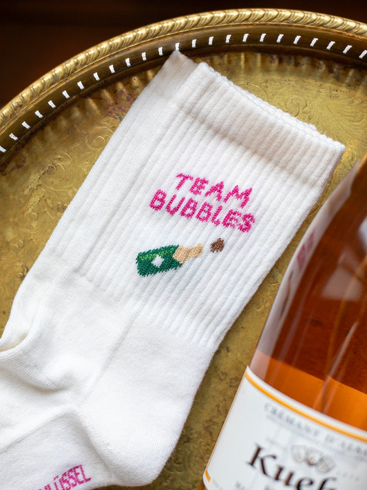 Weisse Sportsocken mit dem pinken Schriftzug «Team Bubbles» und darunter einer kleinen Abbildung einer Proseccoflasche. Darunter ist der Anschnitt eines goldenen Tabletts zu sehen, rechts eine orange Flasche Prosecco.