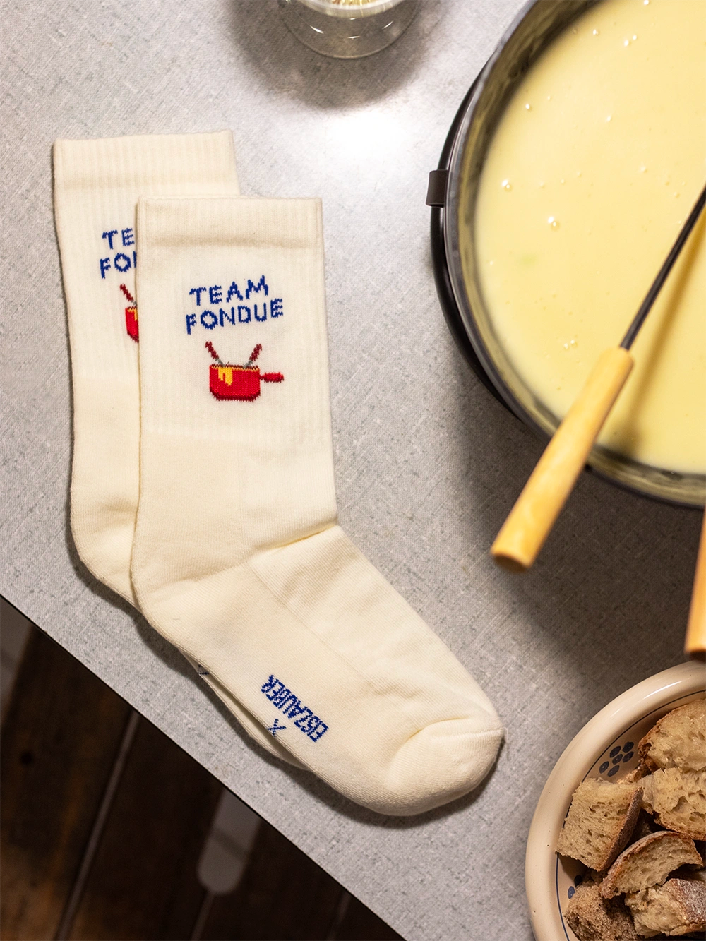 Aufsicht auf einen Tisch mit weissen Sportsocken mit dem Schriftzug Team Fondue. Rechts davon ein angeschnittenes Caquelon mit Fondue und einer Gabel darin, unten geschnittene Brotwürfel