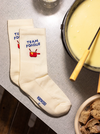 Aufsicht auf einen Tisch mit weissen Sportsocken mit dem Schriftzug Team Fondue. Rechts davon ein angeschnittenes Caquelon mit Fondue und einer Gabel darin, unten geschnittene Brotwürfel