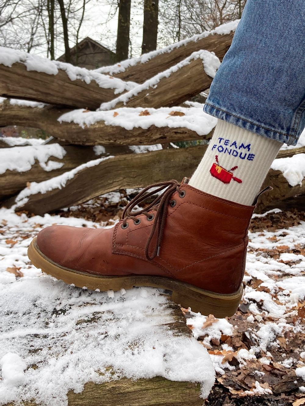 Detailausschnitt von Füssen die weisse Sportsocken mit dem Schriftzug Team Fondue tragen und braune Lederschuhe tragen. Im Hintergrund verschneite Baumstämme.