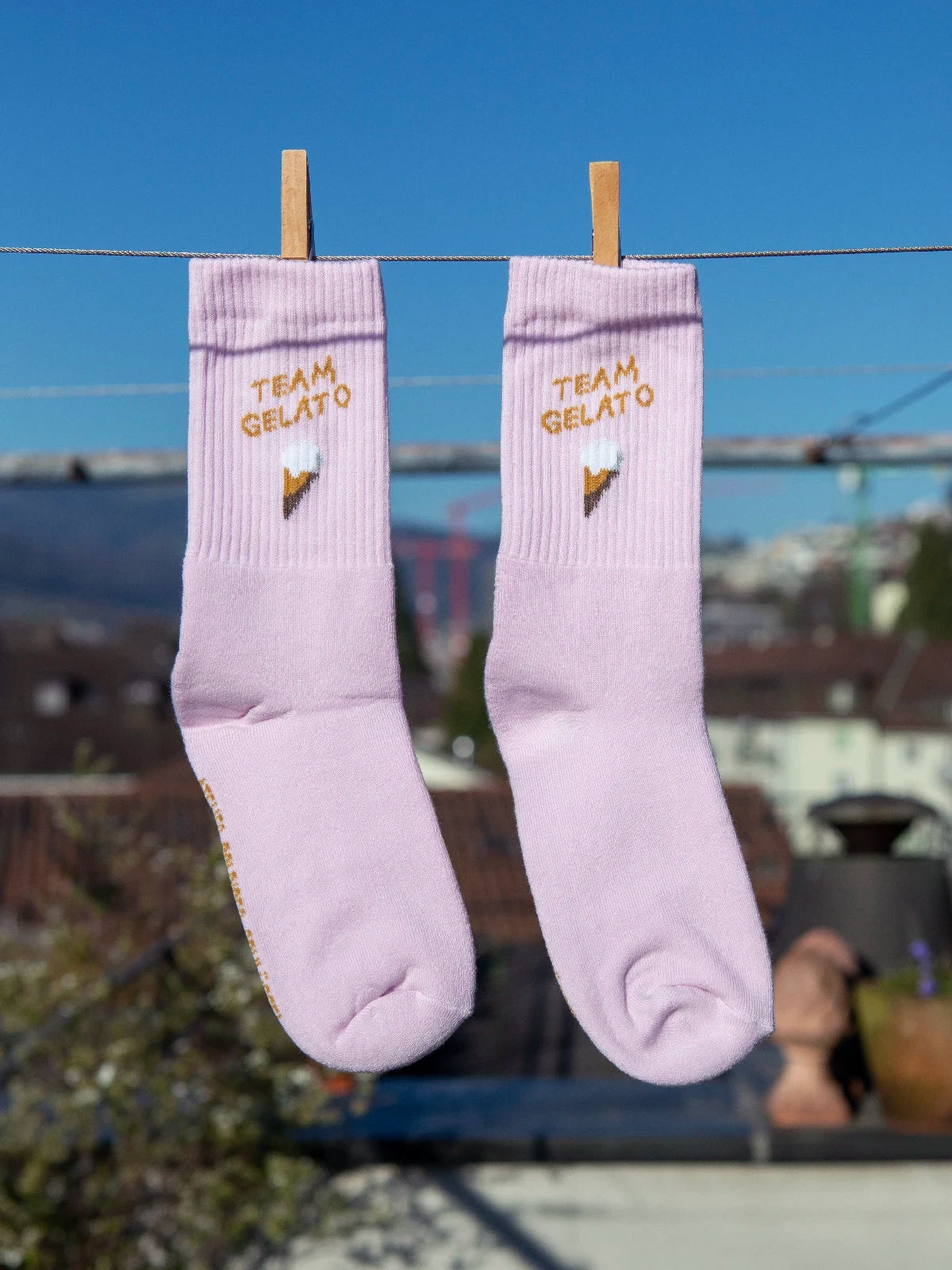 Zwei rosa farbenen Sportsocken hängen an einer Wäscheleine. Im Hintergrund sieht man unscharf Häuser, Hügel und einen blauen Himmel. Im Bündchen der beiden Socken ist der Schriftzug Team Gelato sowie ein Eis in der Waffel gestrickt.