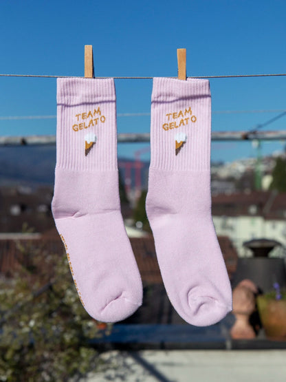 Zwei rosa farbenen Sportsocken hängen an einer Wäscheleine. Im Hintergrund sieht man unscharf Häuser, Hügel und einen blauen Himmel. Im Bündchen der beiden Socken ist der Schriftzug Team Gelato sowie ein Eis in der Waffel gestrickt.