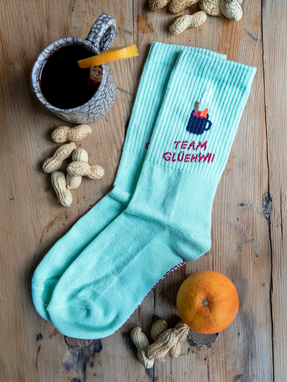 Aufsicht auf mint farbene Socken mit dem Schriftzug Team Glühwein auf holzigem Untergrund. Rund um die Socken liegen Erdnüsse, eine Orange und eine Tasse mit Glühwein.