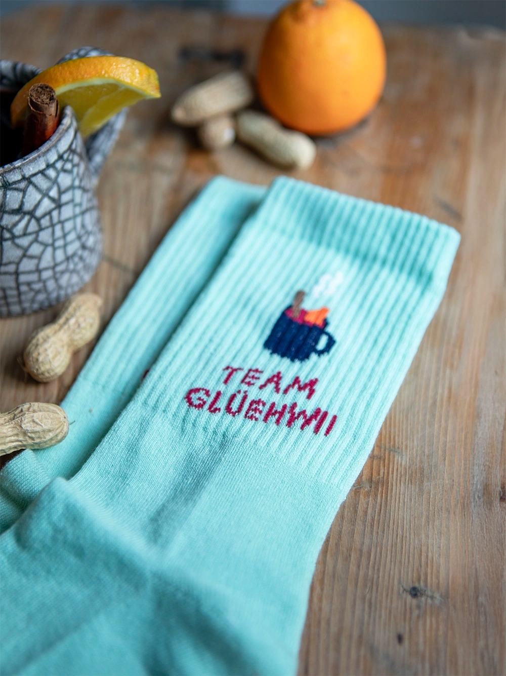 Detailansicht von mint farbenen Socken mit dem Schriftzug Team Glühwein auf holzigem Untergrund. Rund um die Socken liegen Erdnüsse, eine Orange und eine Tasse mit Glühwein.