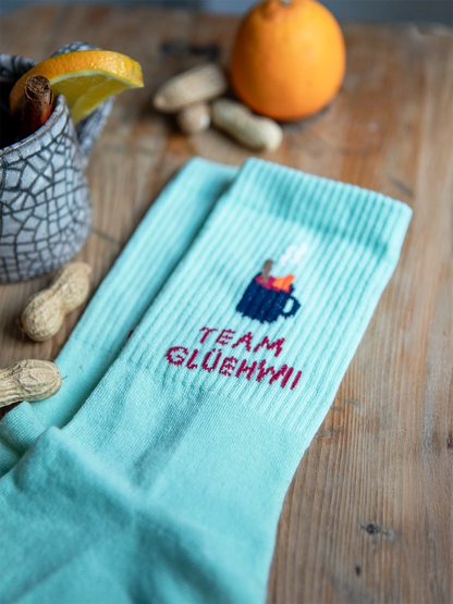 Detailansicht von mint farbenen Socken mit dem Schriftzug Team Glühwein auf holzigem Untergrund. Rund um die Socken liegen Erdnüsse, eine Orange und eine Tasse mit Glühwein.